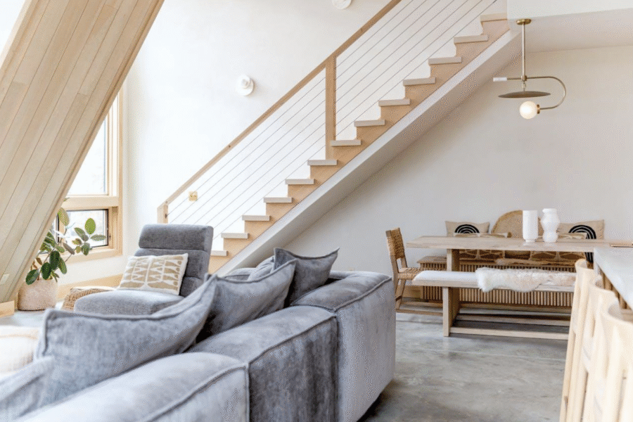 Modern living space with a grey sectional, wood-and-wire staircase, and dining area.